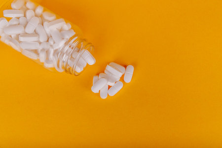Transparent pill bottle with white tablets on a yellow surface, showcasing medical care essentials in a modern styleの写真素材