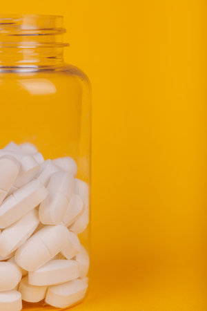 Clear pill bottle with scattered white tablets on yellow, representing healthcare essentials in a bright settingの写真素材
