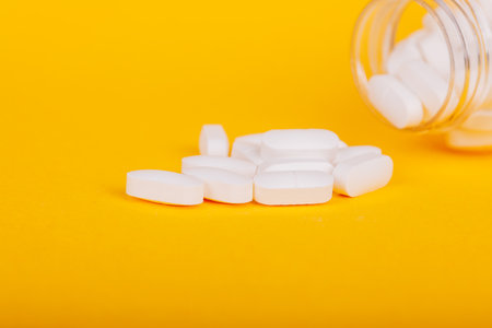 Transparent pill bottle with white tablets scattered on yellow, symbolizing wellness and healthcare