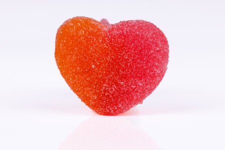 Close-up of a peach-flavored heart gummy on a white background with a wooden spoon full of sugar beside it, all arranged on a wooden tableの写真素材