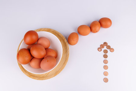 A white bowl filled with brown eggs sits on a wooden board, alongside a row of eggs and a copper coin arrow, highlighting rising egg costsの写真素材