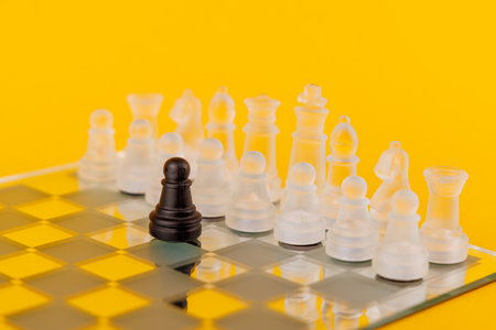 Black pawn standing alone against white pieces on a mirrored chessboard, symbolizing challenge and strategy, with a yellow backgroundの写真素材
