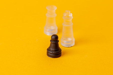 Black pawn stands against white king and queen on yellow chessboard, illustrating strategy, power struggle, and contrast in leadership and decision makingの写真素材