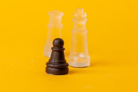 Black pawn opposing white king and queen on yellow background, representing leadership, strategy, and competition. A metaphor for resilience and challengeの写真素材