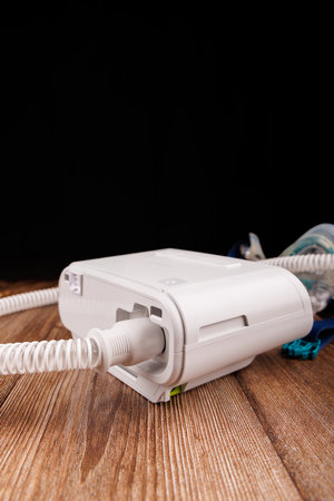 The photo depicts a white CPAP machine with its hose and mask, placed on a dark wooden table,...