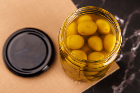 A hand picks an olive from a glass jar using a toothpick, placed on black marble table with a dark backgroundの写真素材