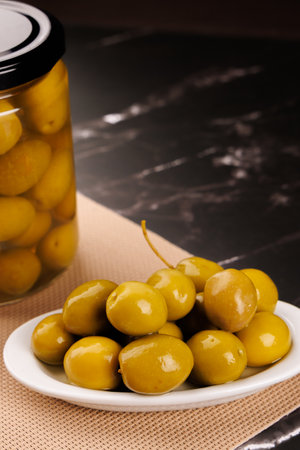 A jar packed with olives next to a serving plate, both on a black marble table with black backgroundの写真素材