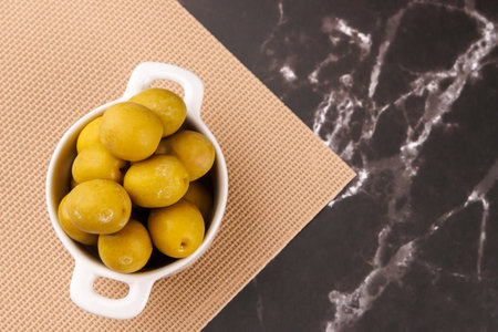 A hand holds a toothpick picking an olive from a bowl next to an oil bottle on black marble, viewed from aboveの写真素材