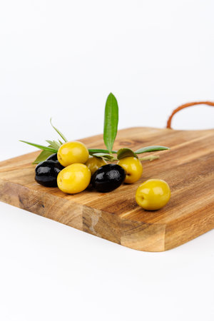 Green and black olives arranged on a dark cutting board with olive branches, offering a rustic Mediterranean touch against a white baseの写真素材