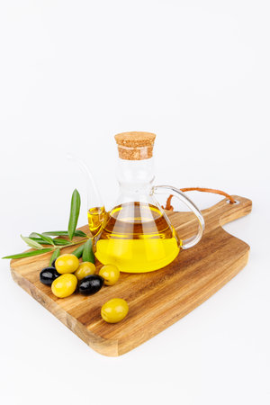 Glass cruet with golden olive oil, surrounded by green olives and olive branches, on a rustic board with white background.の写真素材