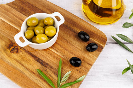 Olive oil bottle with olives and olive branches arranged on a dark chopping board over a white surface.の写真素材