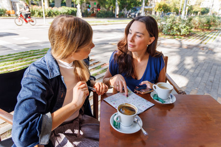Two women relax on a terrace, drink coffee, and share travel experiences in the city.の写真素材