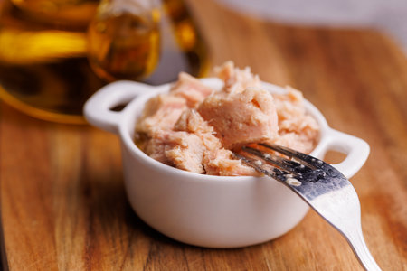 Open metal can of tuna with golden oil placed over a wooden surface in daylight.の写真素材