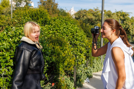 Two friends laugh and take retro style photos with a classic camera as they walk through a bright...