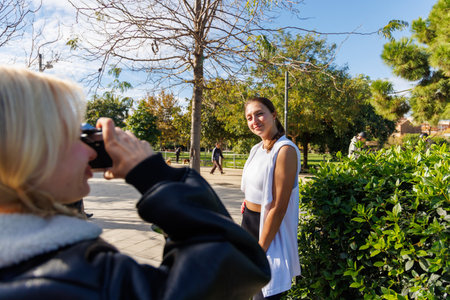Two women walk through a quiet park and take playful photos with a vintage camera, enjoying a fun...