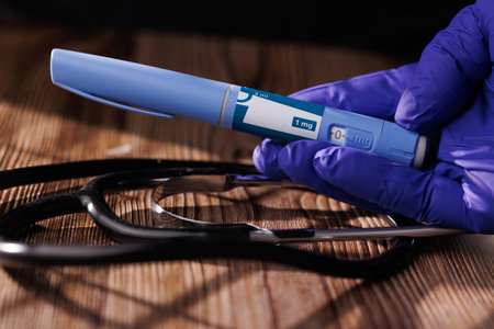Gloved hand with GLP-1 pens and a stethoscope on a dark wooden table represents professional...