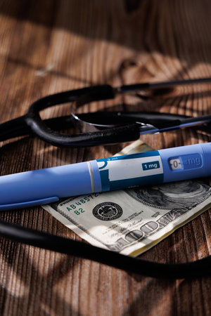 A conceptual shot of GLP-1 pens and a medical stethoscope on a 100 dollar bill over dark woodの写真素材