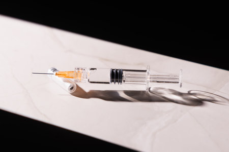 Aesthetic medical shot of a glass syringe on marble under focused light rays against a dark background.の写真素材