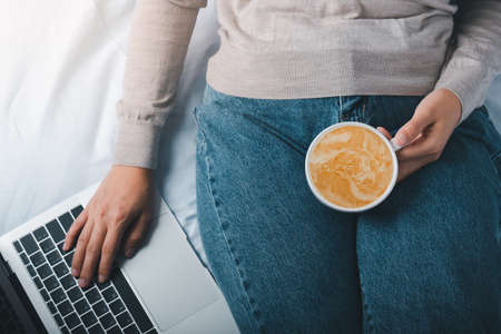 Women hands holding cup of coffee latte at home and checking her laptop.の写真素材