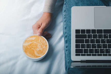 Women hands holding cup of coffee latte at home and checking her laptop.