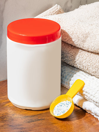 White plastic washing powder container with red cap staying on bathroom table near fresh towels and yellow spoonの写真素材