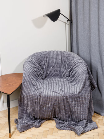 There is a cozy chair that is beautifully covered in a soft, grey blanket, positioned conveniently next to a stylish lamp providing lightの写真素材