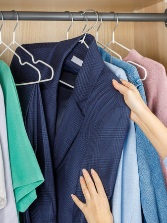 A person is extending their arm to reach for a blue shirt that is hanging in a closet full of various clothing items and accessoriesの写真素材
