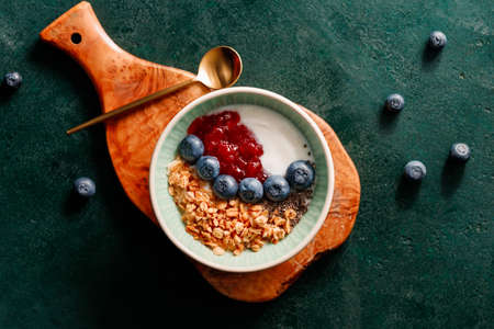 Top view of breakfast with granola, yoghurt, strawberry jam, chia seeds and blueberry on a dark...
