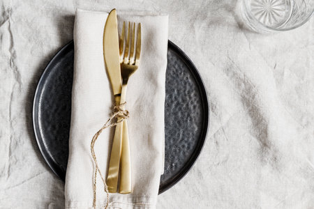 Golden cutlery on a linen napkin with black plate. Decorated table setting for dinner.