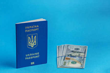 a pack of 100 dollars banknotes lies next to a standing Ukrainian passport one passport on a blue background close-up traveling businessの写真素材