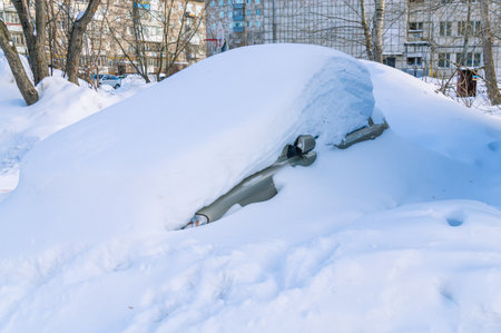In winter, the car is covered with snow. The car is under a snowdrift. An abandoned old car in the yard in winter. Winter in the city. The fallen snow filled up the car. Snow drift in a blizzard.の写真素材