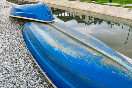 Blue pleasure boats lie upside down on gray pebbles on the shore of a water channel. Walking on the water. The battered bottom of the boat.の写真素材