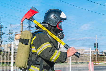 A firefighter in a helmet and breathing apparatus put an assault ax on his shoulder. A firefighter in protective clothing and a mask on the side. A lifeguard with a fire axe. Rescue service.の写真素材