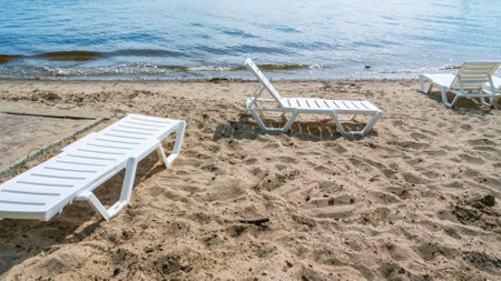 A beach by the river with plastic sun loungers. Sandy beach on the shore. Beach holidays in summer. White plastic sun lounger for sunbathing. vacation at sea.の写真素材
