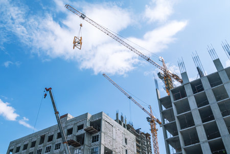 A tower construction crane on the background of a blue sky with clouds. The frame of a building under construction. Building construction. A crane lifts a metal building structureの写真素材