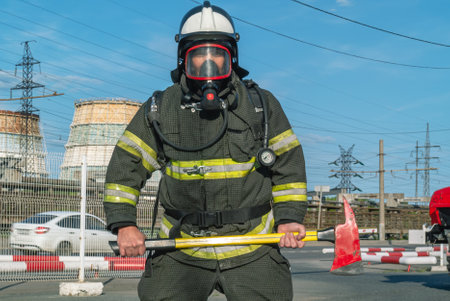 A firefighter in protective clothing and helmet holds an assault ax in his hands. A firefighter on...