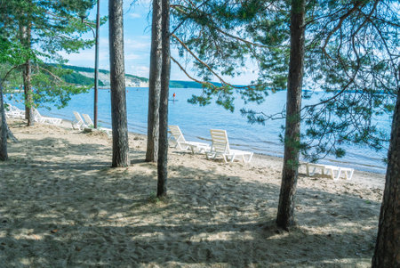 A beach by the river with plastic sun loungers. Sandy beach on the shore. Beach holidays in summer. White plastic sun lounger for sunbathing. Vacation at sea.の写真素材