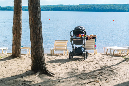 A baby stroller with a child on the beach. A walk with a small child in the fresh air. A baby carriage with a small child stands in the shade of trees near the river. Walking with small children.の写真素材