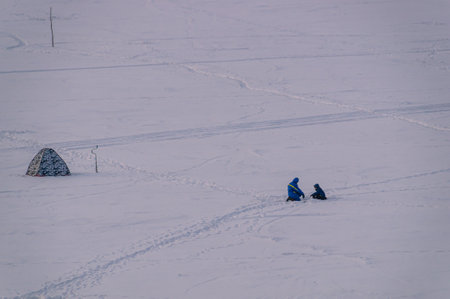 Fishing in winter on the river through a hole in the ice. A wide river covered with ice and snow. Fishermen are fishing on the ice. Anglers use a fishing ice drill to drill holes. Winter on the river.の写真素材