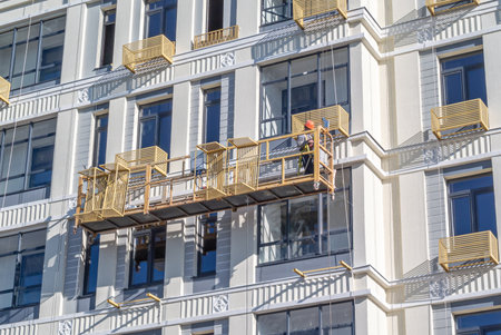 The installer installs baskets for the outdoor air conditioner unit on the facade of the building. The builder in the cradle works at a height. Cladding construction works at height.の写真素材