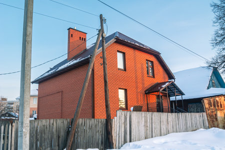 A newly built two-storey red brick house in the village in winter. Snowdrifts near a wooden fence. The cottage is built of brick. An old wooden pole of a power line.の写真素材