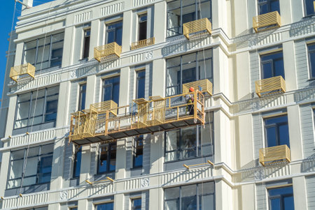 The installer installs baskets for the outdoor air conditioner unit on the facade of the building. The builder in the cradle works at a height. Cladding construction works at height.の写真素材