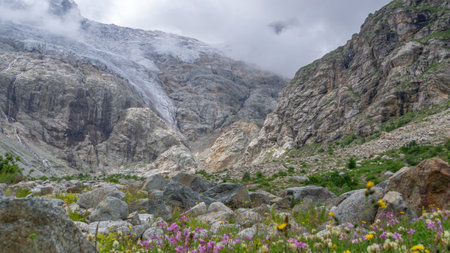 View of a glacier covered with clouds. Panorama in a mountain gorge. The glacier descends from the top of the mountain, forming waterfalls with clear water. Granite stones and flowers in the mountainsの写真素材