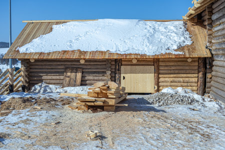 There is snow on the roof of the house. Renovated rustic house built in the 19th century. Cracked dark logs of a wooden house. Flat boards near the house in the countryside.の写真素材