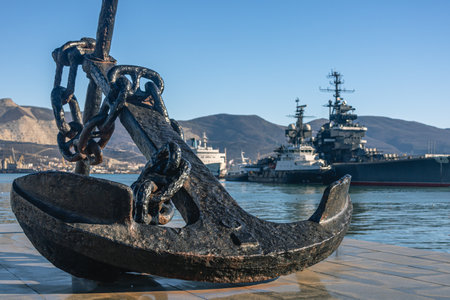 A naval anchor and a warship in the background. Anchor on the Novorossiysk embankment. The marine symbol. Anchor and cruiser in the port.の写真素材