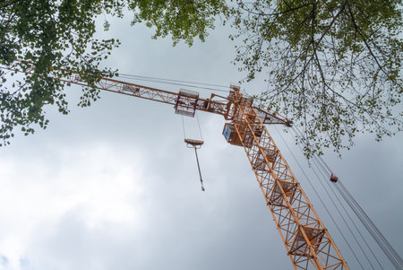 Equipment for the construction and lifting of bulky goods with construction materials. A tower crane on a construction site against a rainy sky. A construction high-rise crane and leaves on a tree.の写真素材
