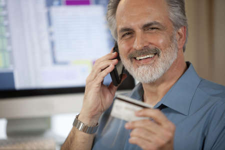 A businessman holding up a credit card and making a payment on his cellphone with a computer monitor in the background. Horizontal shot.の写真素材