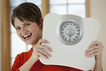 Attractive young woman holds a bathroom scale up while smiling at the camera. Horizontal shot.