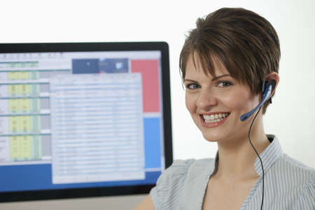 An attractive young businesswoman is wearing a headset and smiling at the camera with a computer monitor in the background. Horizontal shot.の写真素材