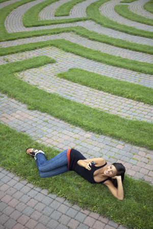 Beautiful young Asian woman smiles as she texts on her cellphone while lying on a grass labyrinth. Vertical shotの写真素材